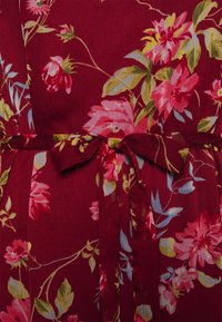 Vestido floral maroon com um laço na cintura. Apresenta flores rosas, amarelas e azuis com folhas verdes sobre um tecido texturizado.