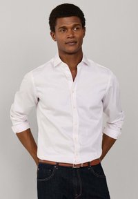 Mand iført en lyserød, stribet skjorte med knapper, ærmerne rullet op til albuerne, mørke jeans og et brunt bælte, stående foran en ensfarvet baggrund.