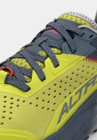Chaussure de course vert lime avec des accents gris, tige en mesh, design perforé et lacets plats gris. Présente un détail matériel rouge.