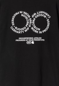 Camiseta negra con texto blanco en alineación circular, incluyendo las frases "RUGGED," "WORK IN PROGRESS," y "GROUNDWORKS." Material de algodón.