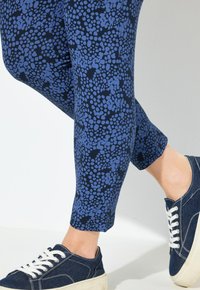 Gambe in pantaloni blu navy con macchie irregolari azzurro chiaro, abbinate a sneakers in denim scuro con lacci e suole bianche su uno sfondo chiaro.