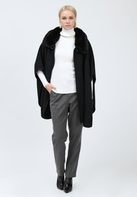 Zwarte wollen cape met een nep-fur kraag, gedragen over een witte geribbelde turtleneck en grijze broek, gecombineerd met zwarte puntige enkellaarsjes.