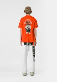 Camisa roja de manga corta con un gráfico de un caniche vestido con una chaqueta blanca. Combinada con pantalones blancos de corte holgado y calzado gris.
