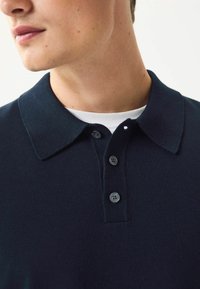 Tumes poloshirt tekstureeritud kraega, kolme nööbiga klapp ja kihiline valge sissekandja, mis on nähtav all. Sile kangasviimistlus.