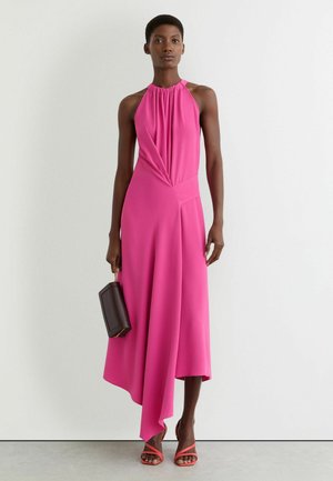 Vrouw draagt een mouwloze felroze asymmetrische jurk, houdt een donkerbruine clutch vast en draagt koraalkleurige sandalen met bandjes.