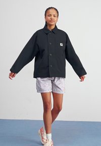 Zwarte oversized jas met een kraag en knopen aan de voorkant, voorzien van een klein logo zakje. Gecombineerd met gestreepte shorts en sneakers.