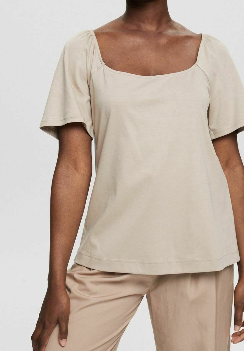 Femme portant un haut beige à manches courtes avec un col carré et un pantalon beige assorti sur un fond clair uni.