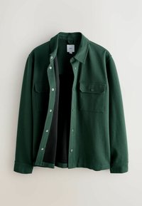 Donkergroen overhemd van gestructureerde stof, met twee borstzakken en een kraag, open gedragen om een zwarte voering zichtbaar te maken.