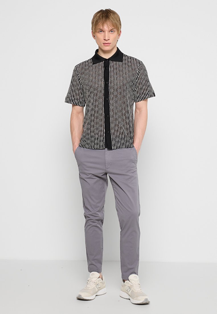 Młody mężczyzna stoi z rękami w kieszeniach, ubrany w koszulę z krótkim rękawem w czarno-białe wzory, szare spodnie i beżowe sneakersy.
