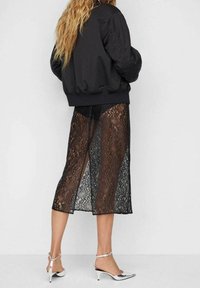 Veste bomber noire avec poignets côtelés, associée à une jupe midi en dentelle noire transparente avec des fentes, portée avec des chaussures à talons argentés.