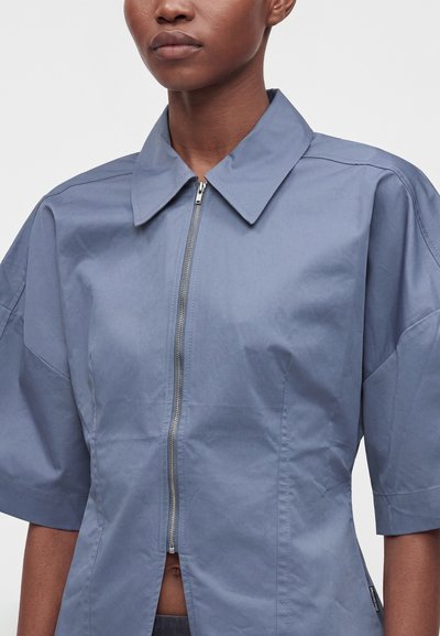 Femme portant une chemise bleu clair à manches courtes avec une fermeture éclair argentée sur le devant et un large col pointu, raccourcie à la taille.