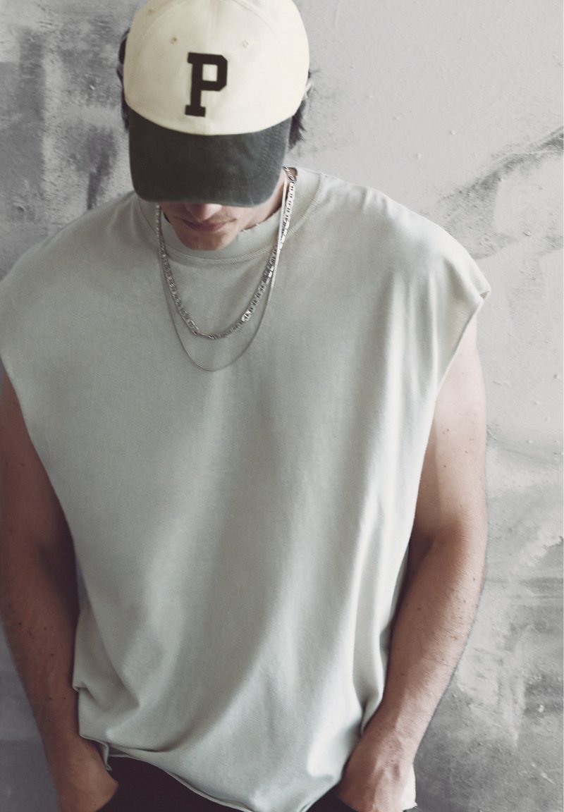 Homme portant un débardeur beige, deux chaînes argentées et une casquette noir et blanc avec la lettre P, regardant vers le bas devant un mur texturé.