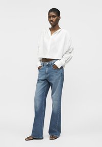 Femme portant un chemisier blanc court, un jean bleu à jambes larges et des sandales noires, debout les mains dans les poches devant un fond uni.