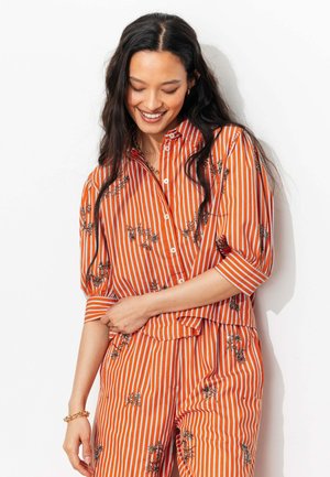 Femme aux longs cheveux foncés portant un chemisier à fleurs rayé orange et blanc et un pantalon assorti, souriante et regardant vers le bas sur fond blanc.