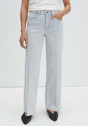 Persoon die lichtgrijze jeans met wijde pijpen en zwarte puntige schoenen draagt, met één hand in de zak en een wit shirt.