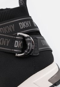 Baskets noires avec sangle élastique marquée DKNY et boucle métallique argentée au-dessus d'une semelle blanche sur fond blanc.