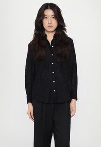 Zwarte button-up shirt van doorschijnende stof, met lange mouwen en een afgeronde zoom. Gepaard met losse zwarte broek. Eenvoudige witte knopen.