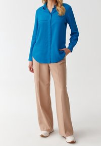 Blå langærmet skjorte med knap-ned krave, let buet kant; beige vidde bukser; hvide sneakers med lysebrune såler.