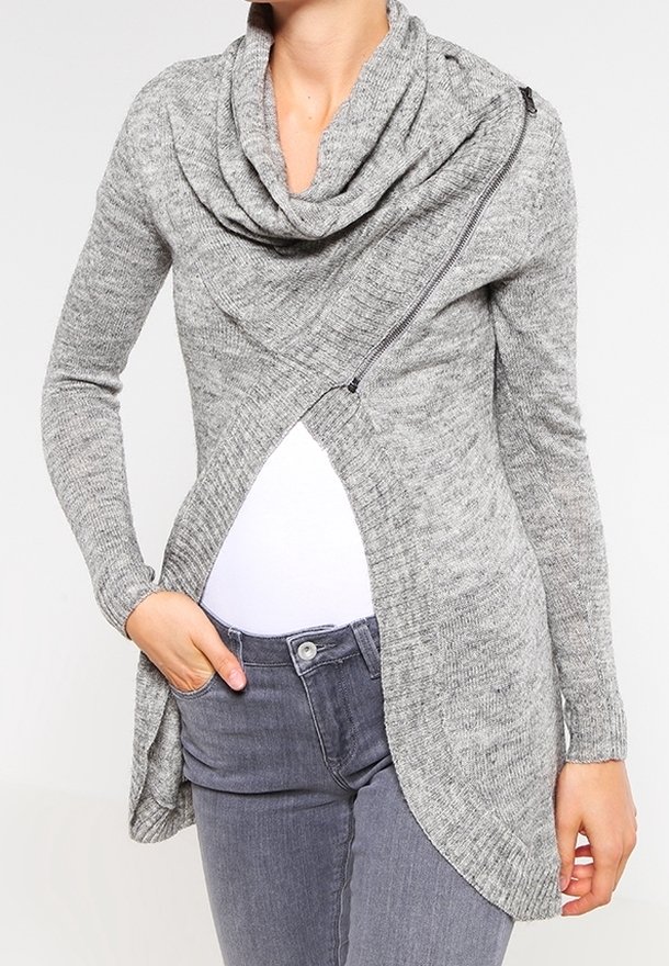 Femme portant un cardigan tricoté asymétrique gris avec un grand col châle et une fermeture éclair diagonale, associé à un haut blanc et un jean gris.