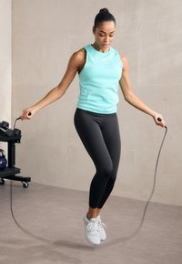 En person som har på seg en lys teal uten ermer topp og svarte leggings hopper tau. De har hvite joggesko og en jevn bakgrunn.