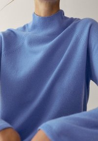 Pull en maille bleu clair à col montant, présentant une texture douce, un col côtelé et des épaules sans couture, avec des manches longues visibles.