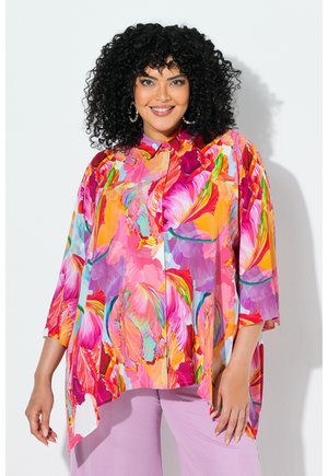 Mujer con cabello rizado y negro sonríe, lleva una camisa grande con estampado floral en tonos rosa, naranja y púrpura sobre pantalones lavanda, frente a una pared blanca.