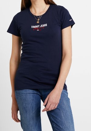 Kvinde iført en marineblå Tommy Jeans t-shirt med rødt og hvidt logo, parret med blå jeans og lagdelte guldkæder.