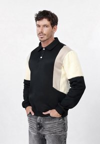 Homme aux cheveux courts et foncés portant un polo à manches longues noir, beige et crème, debout les mains dans les poches de son jean gris.