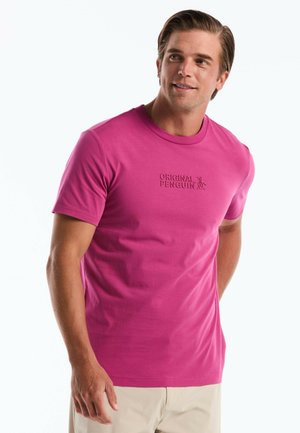 Mann trägt ein leuchtend pinkes T-Shirt mit dem "Original Penguin"-Logo, beige Hose, schaut leicht nach rechts, lächelt sanft vor weißem Hintergrund.