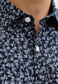 Mustast särgist lähedalt, millel on väikesed valged lillemustrid ja valged nööbid, krae juurest osaliselt lahti nööbitud.