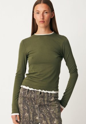 Femme portant un haut à manches longues vert olive avec un bord blanc et un pantalon à motif camouflage, debout devant un fond uni.