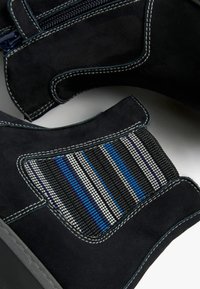 Des bottines en suède noir présentent une fermeture éclair sur le côté, des coutures blanches contrastantes et un panneau en maille rayée bleu, blanc et gris.