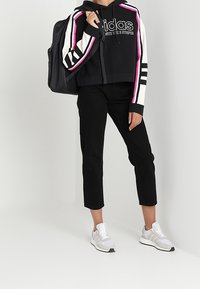 Osoba oblečená v černé mikině Adidas s růžovými a bílými pruhy, černých zkrácených kalhotách, bílých teniskách a nese černý batoh.