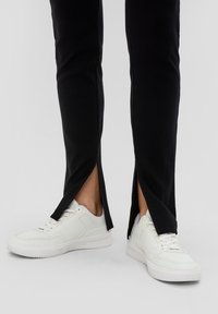 Baskets blanches en cuir texturé, associées à un pantalon noir avec un design à fente sur le côté. Les matériaux sont lisses et ajustés.