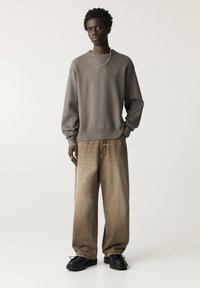 Grauer, oversized Pullover, beige, weit geschnittene Denim-Jeans und schwarze Plateauschuhe; kombiniert mit einer silbernen Kettenhalskette. Insgesamt glatte Textur.