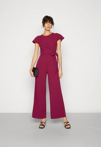 Žena u magenta kombinezonu kratkih rukava širokih nogavica, s vezanom strukom, drži crni clutch i crne sandale s remenčićima, stoji uz bijeli zid.