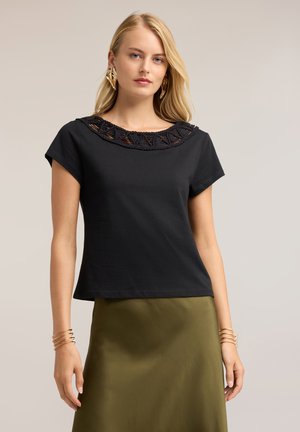Vrouw met recht blond haar die een zwarte top met korte mouwen en kanten halslijn draagt, een olijfgroene rok, gouden blad-oorbellen en armbanden.