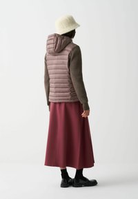 Žena v béžovom vedierkovom klobúku, mauve prešívanej kapucňovej veste, hnedých pletených rukávoch, dlhých marhuľových sukni, čiernych ponožkách a čiernych polobotkách, stojí bokom.