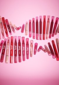 Una colección de productos para labios en varios tonos de rosa, presentados en tubos cilíndricos con acabados transparentes y mate, algunos con detalles brillantes.