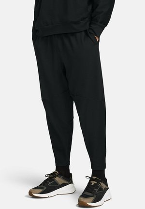 Osoba stojící s rukama v kapsách, oblečená v volných černých kalhotách a černých teniskách s béžovými detaily, na jednobarevném pozadí.