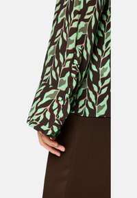 Langärmelige Bluse mit schwarzem Grund, die ein grünes und rotes Blattmuster aufweist, abgerundet mit einem gerafften Bündchen und subtilen quadratischen Akzenten.