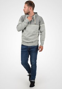 Haut de survêtement en tricot gris avec une rayure horizontale gris clair, capuche avec cordon de serrage ajustable, associé à un jean bleu foncé et des baskets noires.