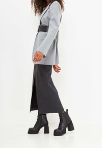 Blazer gris a medida con un cinturón ancho negro, combinado con una falda larga negra a rayas con una abertura lateral y botas negras de tacón grueso.