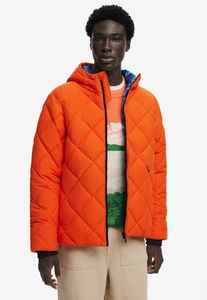 Chaqueta acolchada naranja con capucha, con un forro azul y detalles en negro. Llevada sobre un suéter de patrón multicolor.