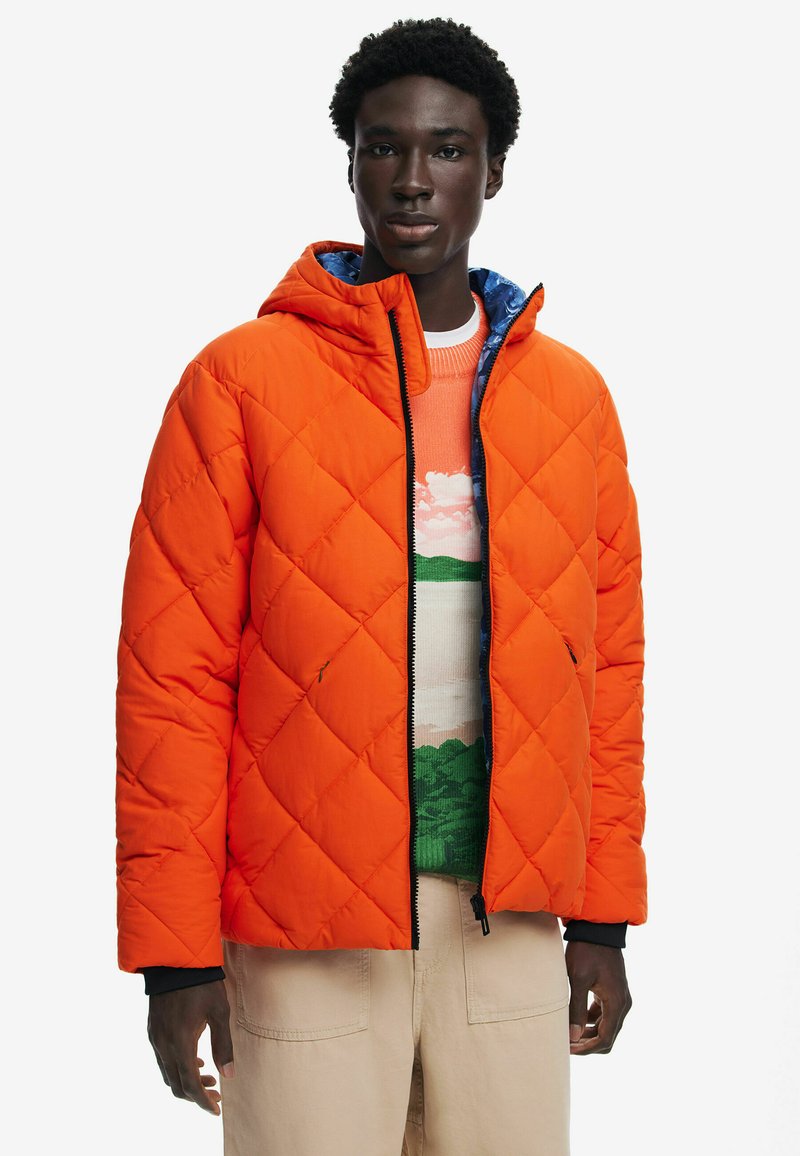 Giacca a vento arancione trapuntata con cappuccio, con rivestimento blu e dettagli neri. Indossata sopra un maglione a fantasia multicolore.