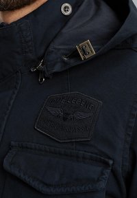 Giacca blu navy in tessuto texturizzato, con cappuccio e una patch nera con il testo "PME Legend American Classic" e grafica di ali.