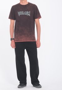 Henkilö, jolla on päällään haalistunut musta Volcom-t-paita, löysät mustat housut, kuluneet mustat kengät ja musta rannekello, seisoo valkoista taustaa vasten.