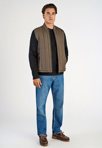 En vadderad väst i tan över en svart ribbad tröja, parat med blå jeans och bruna snörskor. Vesten har dragkedja fram och sidofickor.