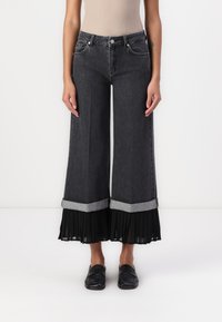 Høy midje, vide ben svarte denimjeans med en kontrasterende grå brettkant og et plissert svart stoffpanel nederst.