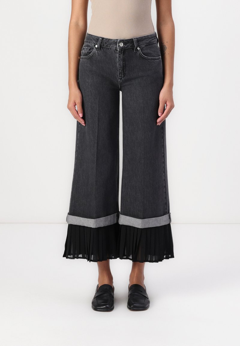 Høy midje, vide ben svarte denimjeans med en kontrasterende grå brettkant og et plissert svart stoffpanel nederst.